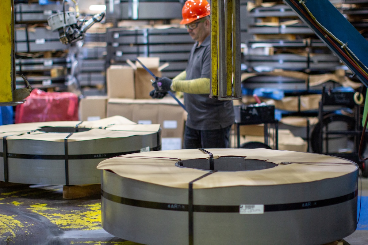 Worker securing large steel coils with metal strapping inside an industrial packaging facility.